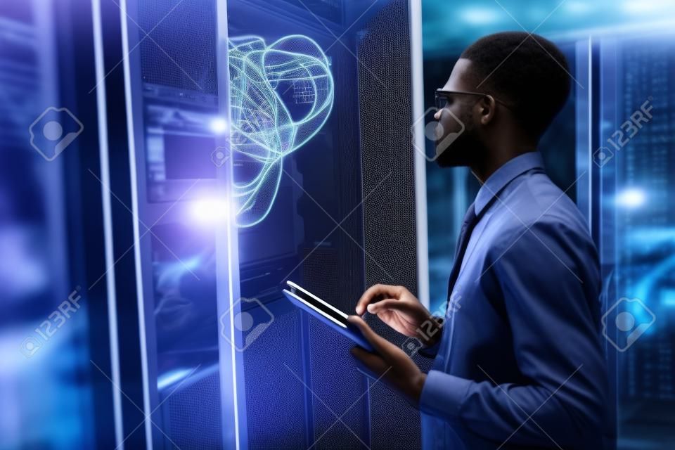 Retrato da vista lateral do homem afro-americano que está pelo armário do servidor ao trabalhar com o supercomputador no centro de dados e segurando o tablet, espaço da cópia