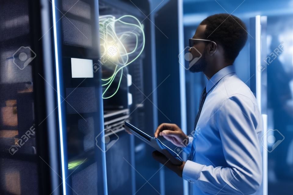 Retrato da vista lateral do homem afro-americano que está pelo armário do servidor ao trabalhar com o supercomputador no centro de dados e segurando o tablet, espaço da cópia