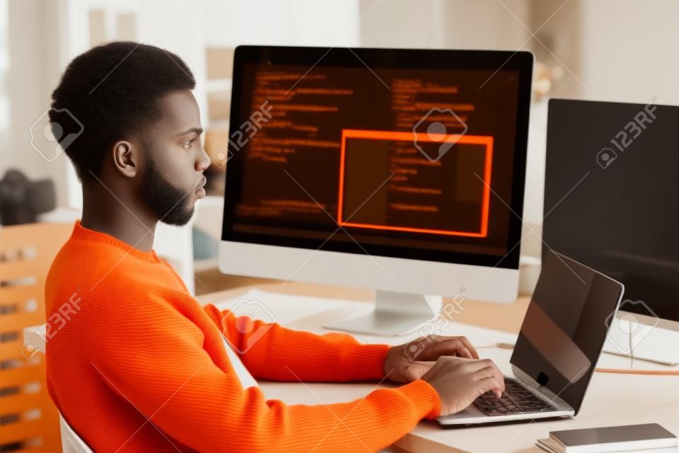 Serious young Black man in warm orange sweater concentrated on coding