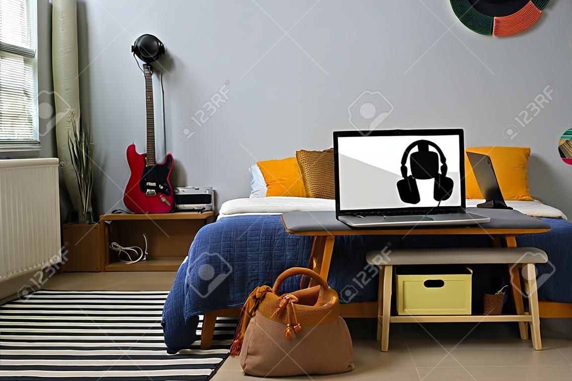 Part of domestic room with comfortable double bed with several cushions and soft bench with headphones hanging on laptop with blank screen