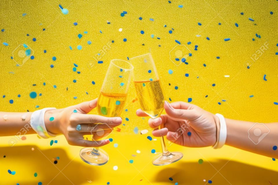 Nahaufnahme von zwei Händen, die Champagnergläser vor einem leuchtend gelben Hintergrund mit Konfetti anstoßen