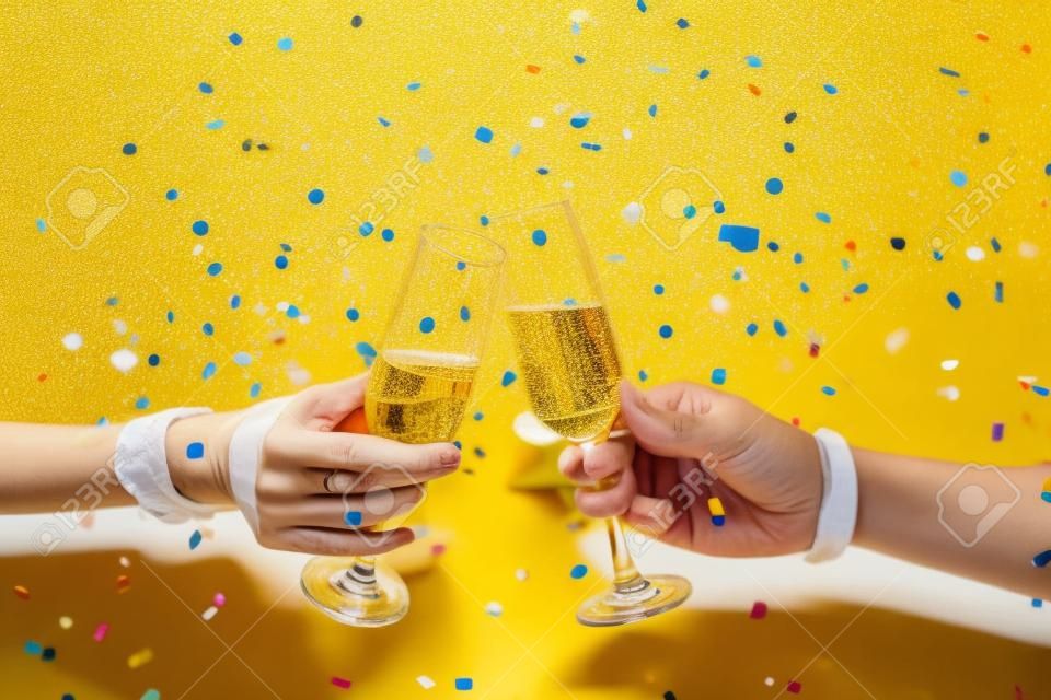 Gros plan de deux mains tintant des verres de champagne sur fond jaune vif avec des confettis