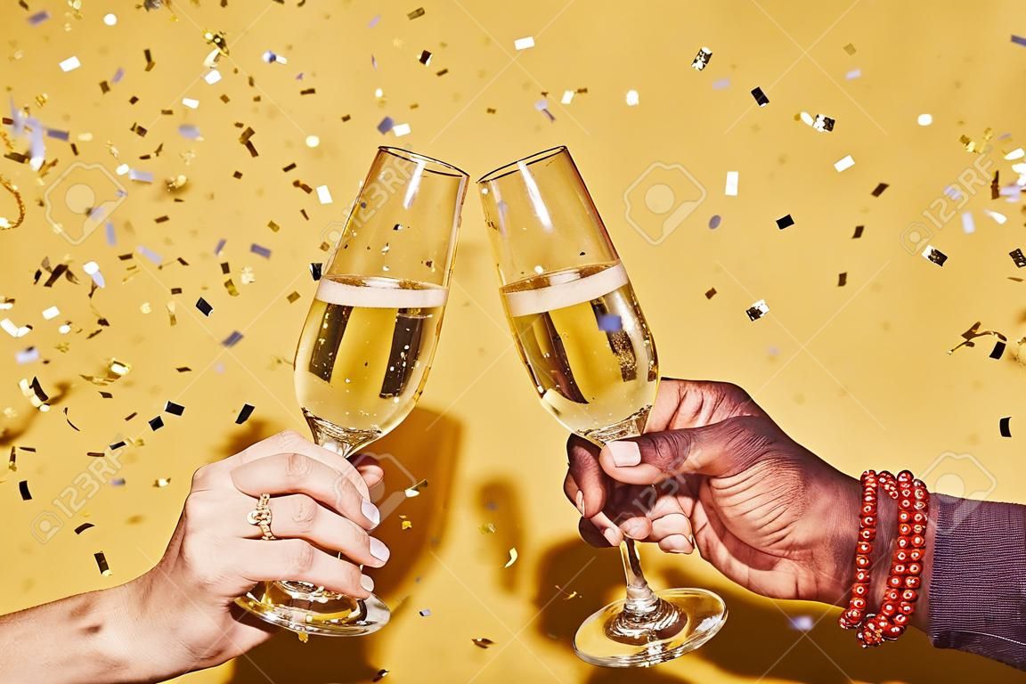 Closeup of two hands clinking champagne glasses against vibrant yellow background with confetti