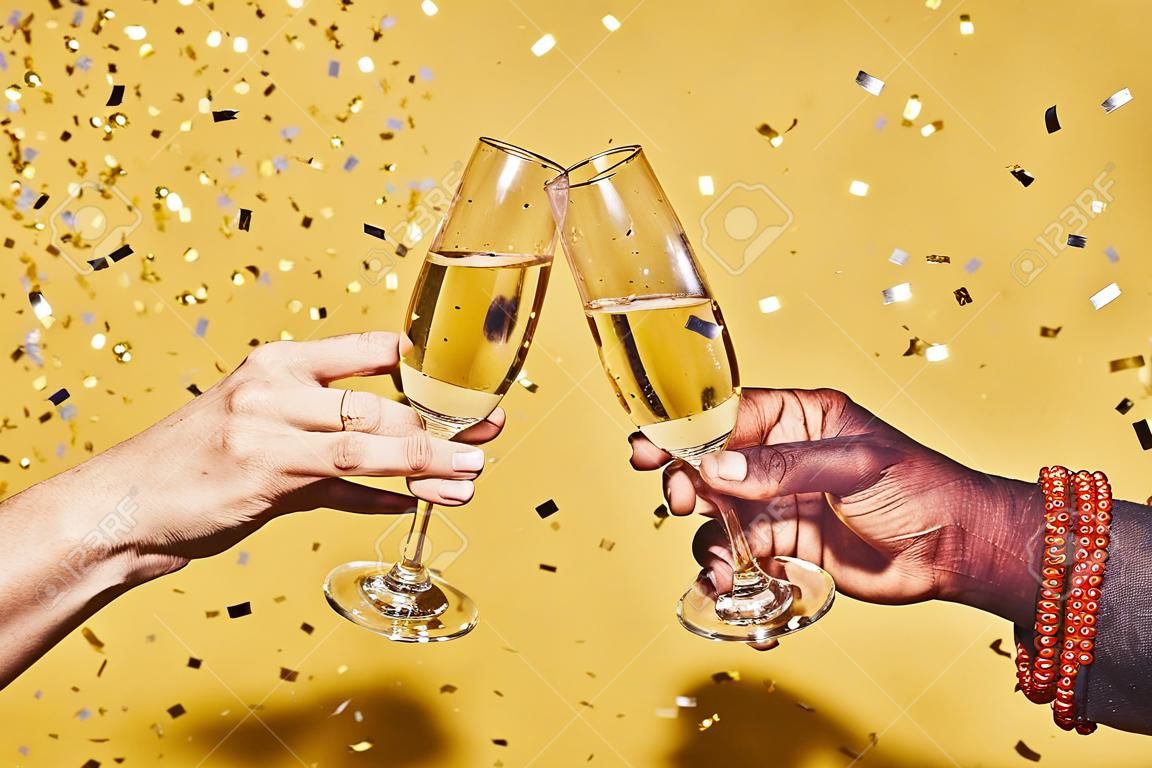 Zbliżenie dwóch rąk brzęczących kieliszkami szampana na żywym żółtym tle z konfetti