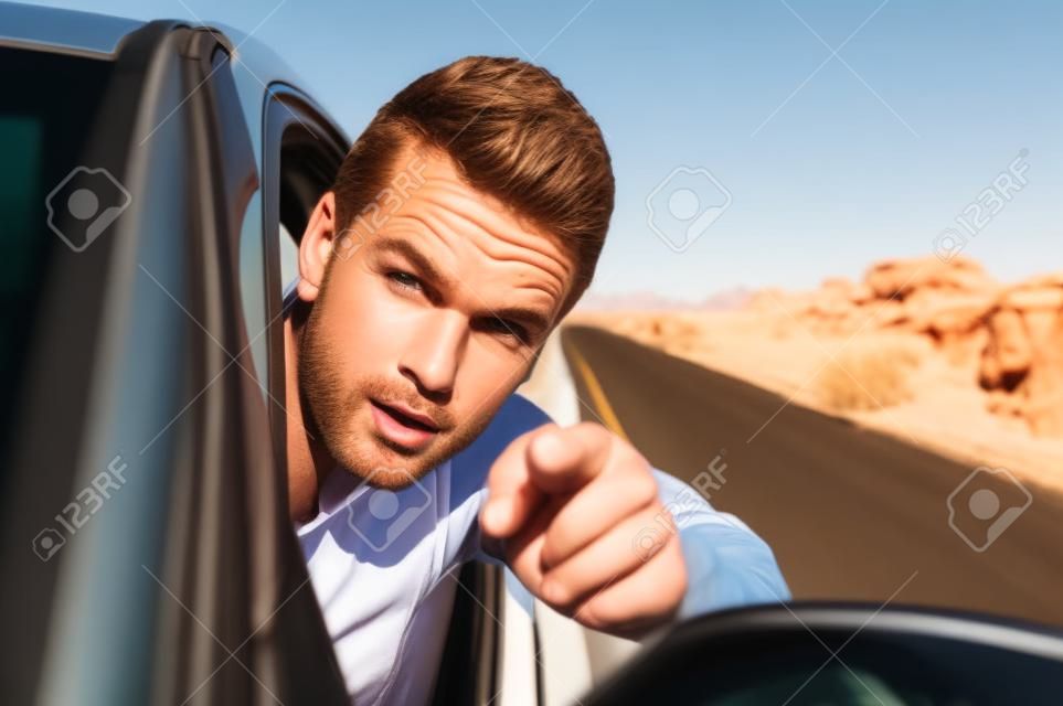 Closeup of handsome free Caucasian man driving car with head out window down desert road as he accusingly points finger at camera