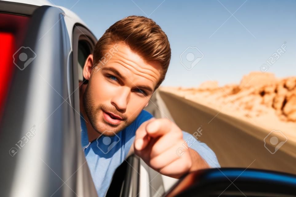Closeup of handsome free Caucasian man driving car with head out window down desert road as he accusingly points finger at camera