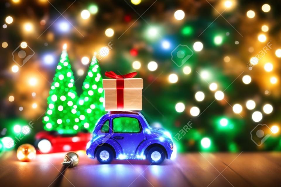 New year decoration and background for greetings with free space for text. Toy car carries a gift on the background of Christmas trees and lights bokeh on a wooden. 2020