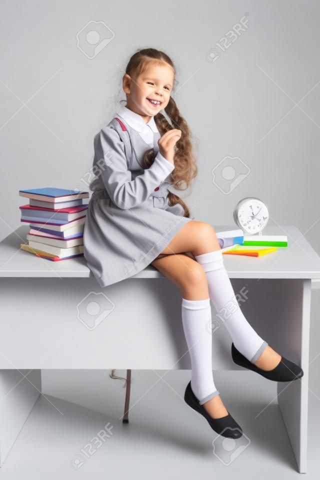 A aluna de fantasia no uniforme da escola senta-se na mesa e sorri alegremente em um fundo cinza claro. De volta à escola. O novo ano letivo. Conceito de educação infantil.