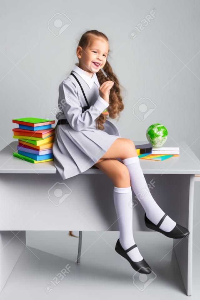 Школьница-непоседа в школьной форме сидит на столе и радостно улыбается на светло-сером фоне. Обратно в школу. Новый учебный год. Концепция детского образования.