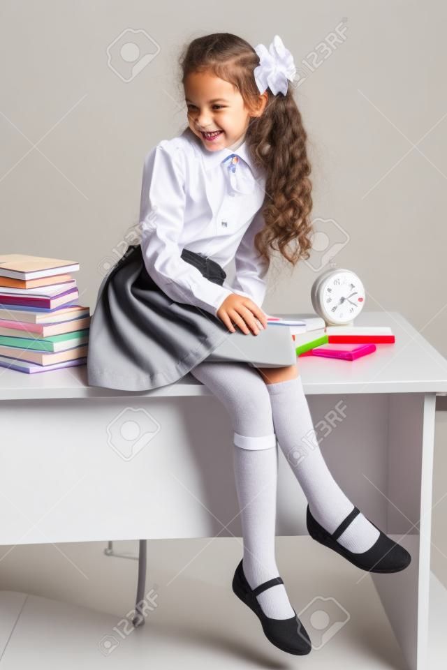 A aluna de fantasia no uniforme da escola senta-se na mesa e sorri alegremente em um fundo cinza claro. De volta à escola. O novo ano letivo. Conceito de educação infantil.
