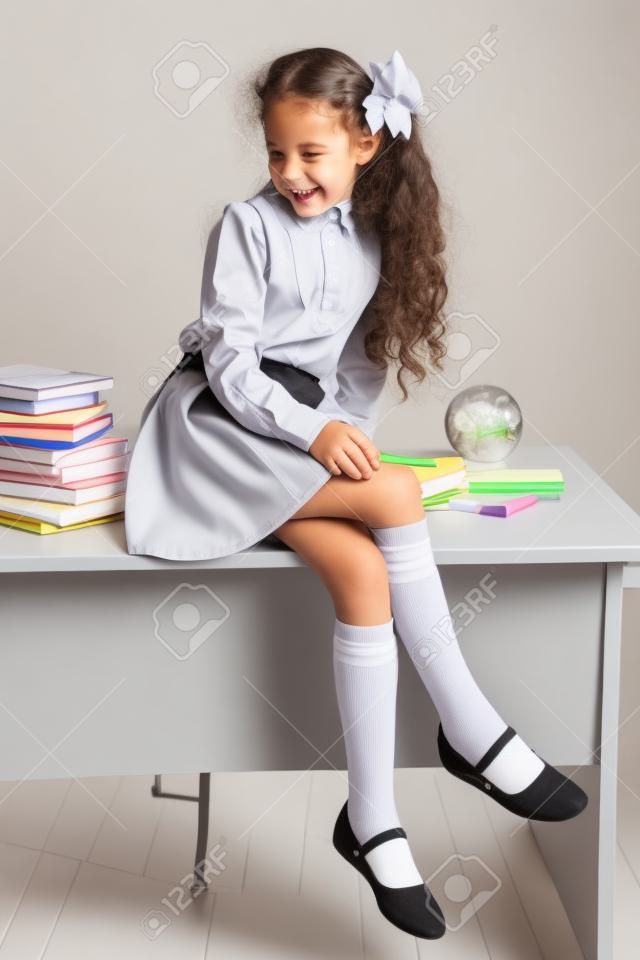 De fidget schoolmeisje in schooluniform zit op tafel en glimlacht gelukkig op een licht grijze achtergrond. Terug naar school. Het nieuwe schooljaar. Kinderonderwijs concept.