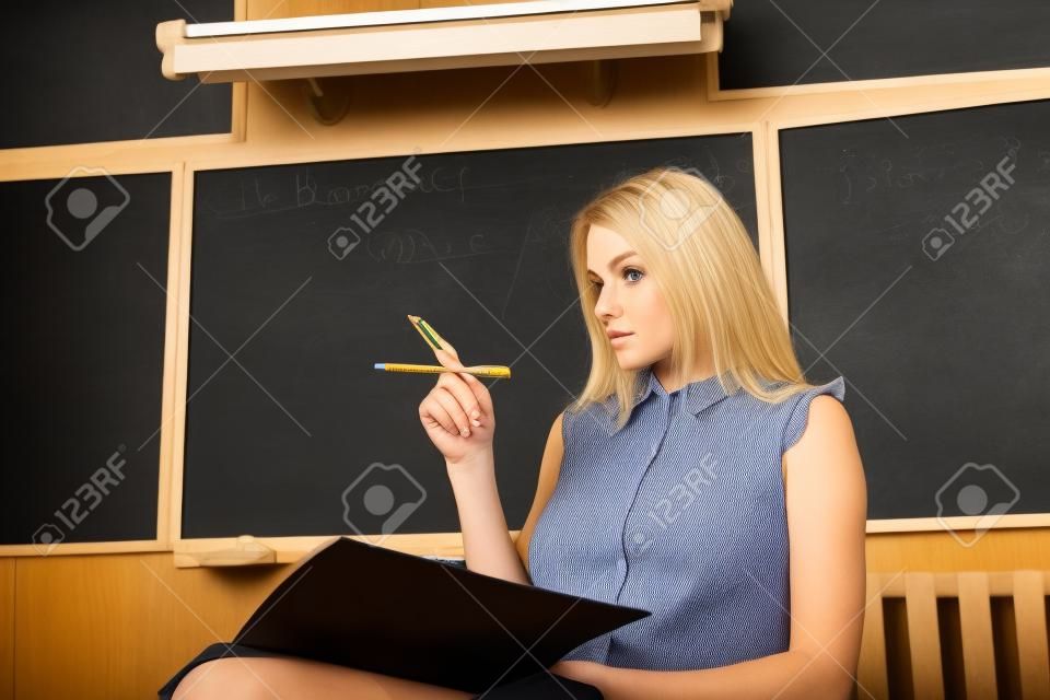 strict jeune professeur blond assis sur une chaise dans la salle de classe et un tableau noir contre l & # 39 ; épaule à un sujet sur les devoirs