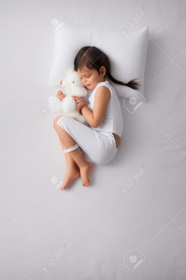 Vista superior de la foto de la niña linda que duerme en la cama blanca con el oso de peluche. Pose tranquila feto. Concepto de posturas para dormir