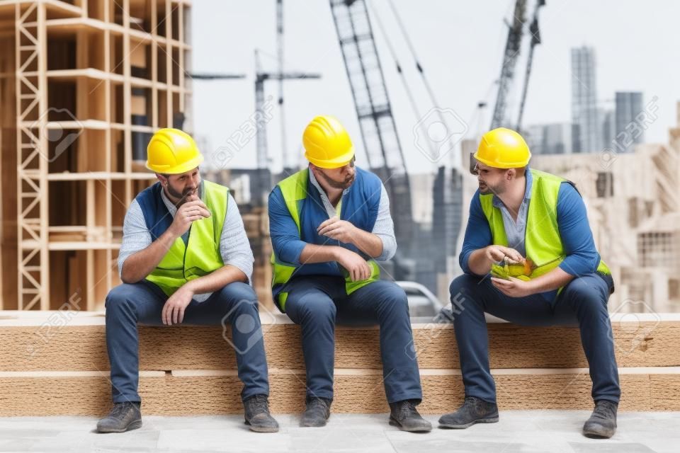 Hora de uma pausa. Grupo de construtores em uniforme de trabalho estão comendo sanduíches e falando enquanto sentado na superfície de pedra contra o canteiro de obras. Conceito de construção. Conceito de almoço
