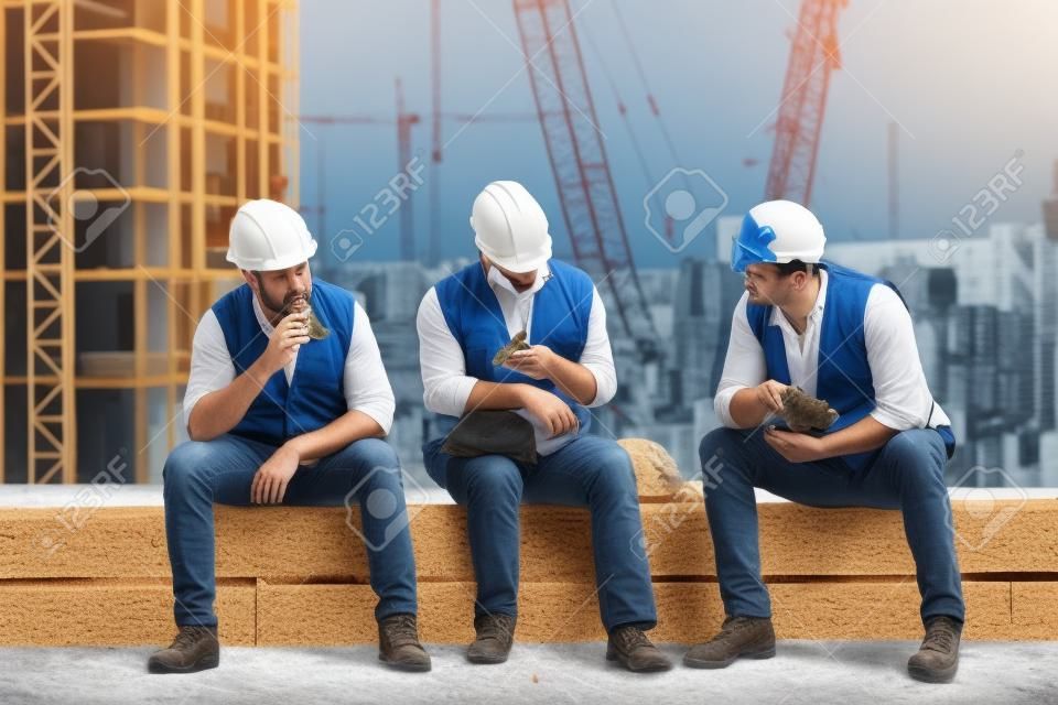 Time for a break. Group of builders in working uniform are eating sandwiches and talking while sitting on stone surface against construction site. Building concept. Lunch concept