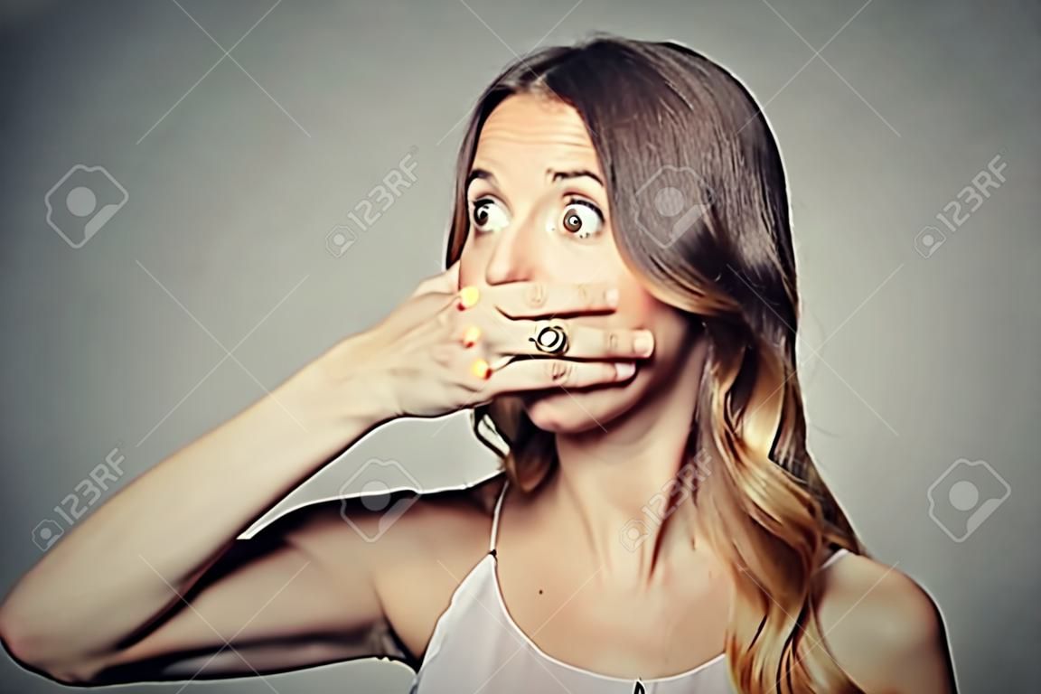 Portrait of scared young woman covering with hand her mouth isolated on gray wall background