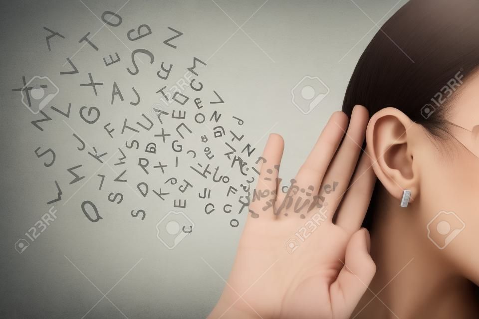 La mujer sostiene su mano cerca de la oreja y escucha cuidadosamente las cartas del alfabeto que vuelan en lugares aislados en fondo de la pared gris