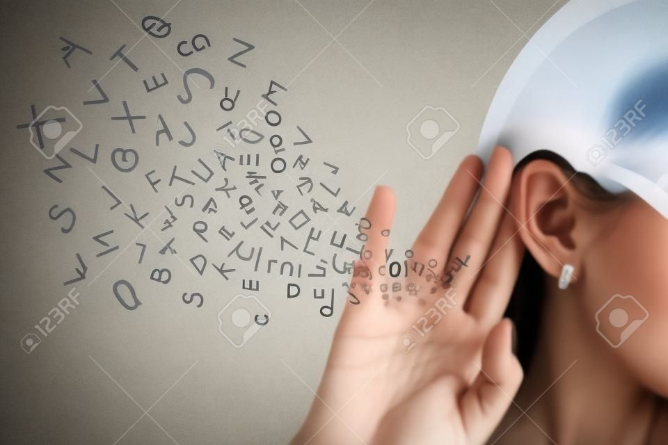 Woman holds her hand near ear and listens carefully alphabet letters flying in isolated on gray wall background 