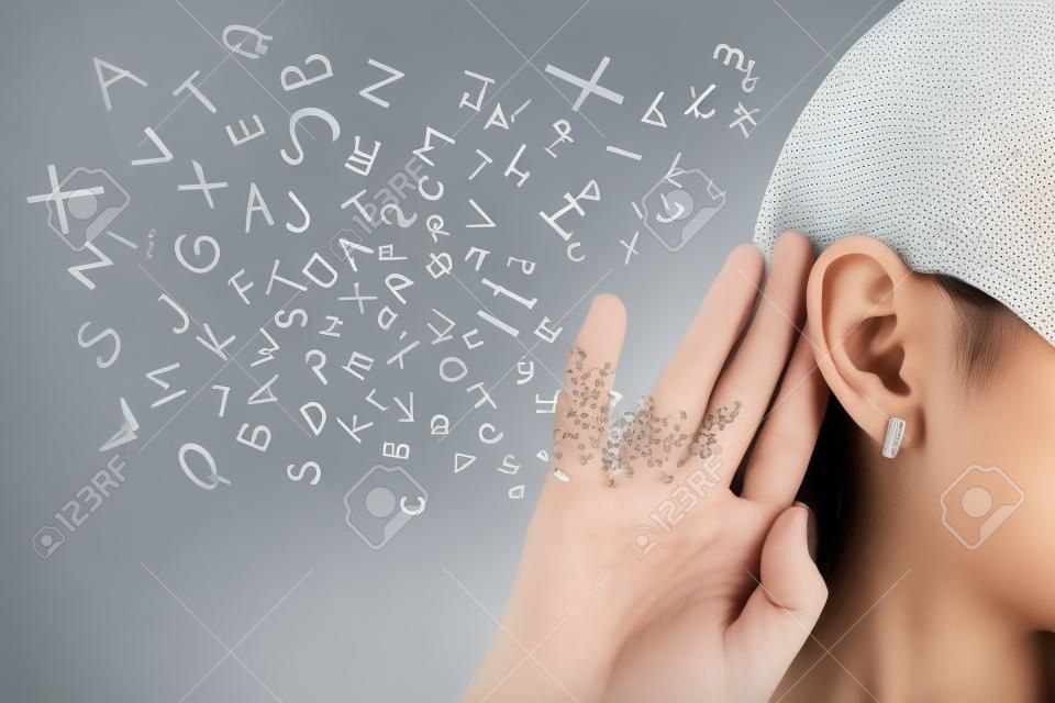 Vrouw houdt haar hand dicht bij het oor en luistert zorgvuldig alfabet letters vliegen in geïsoleerd op grijze muur achtergrond