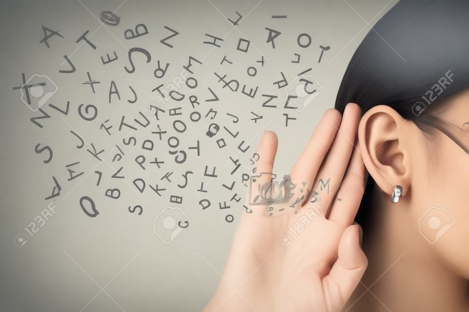 Mulher segura a mão perto da orelha e ouve cuidadosamente letras do alfabeto voando em isolado no fundo da parede cinza