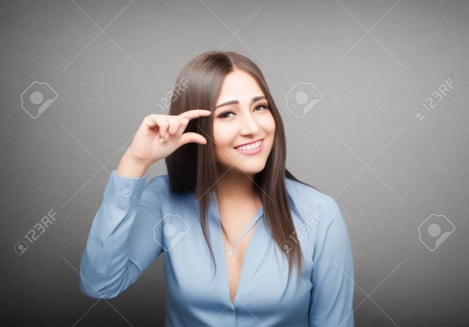 Woman showing small amount size gesture with hand fingers isolated on gray wall background. Human emotion facial expression feelings symbol body language