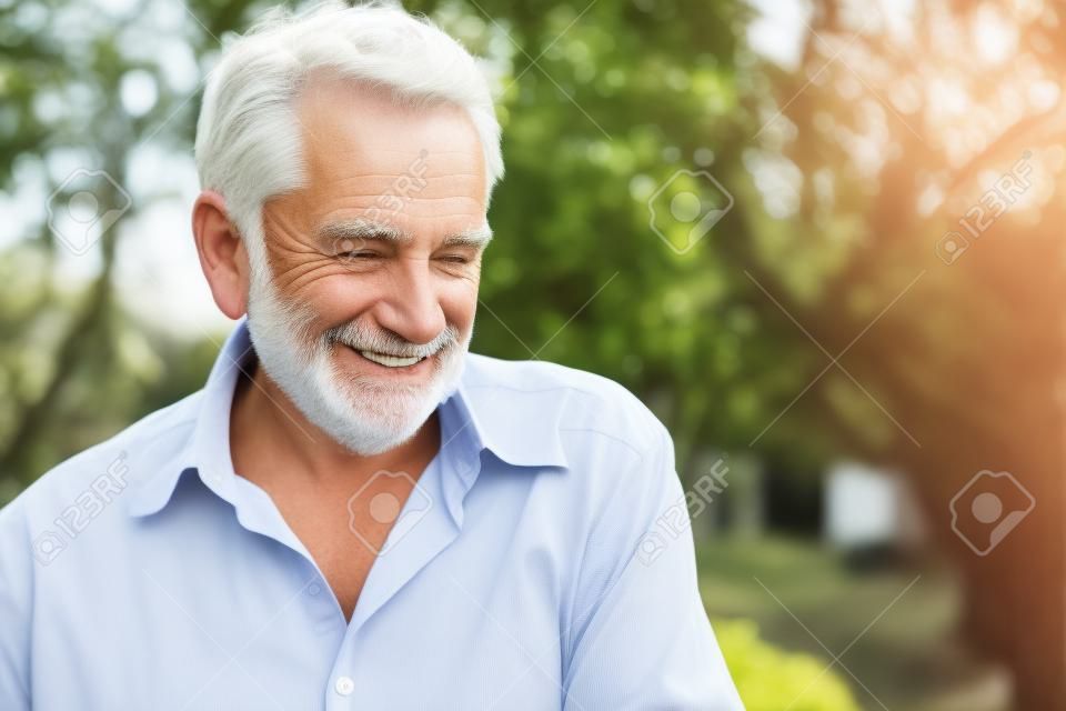 Older white man with grey hair, wearing a shirt, smiling and looking away in outdoor location with natural light.