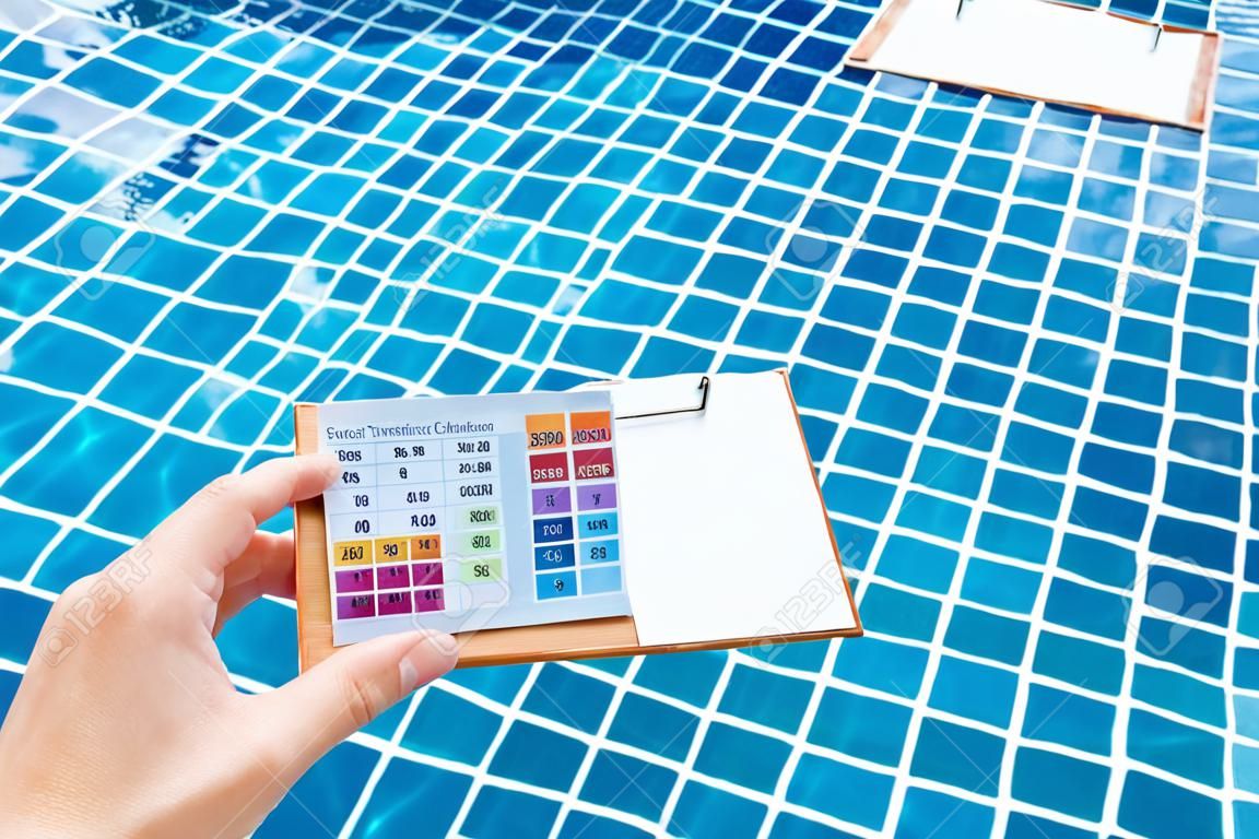 Female hand holding water tester and blank paper on wooden clipboard over clear swimming pool water background, pool water treatment checklist