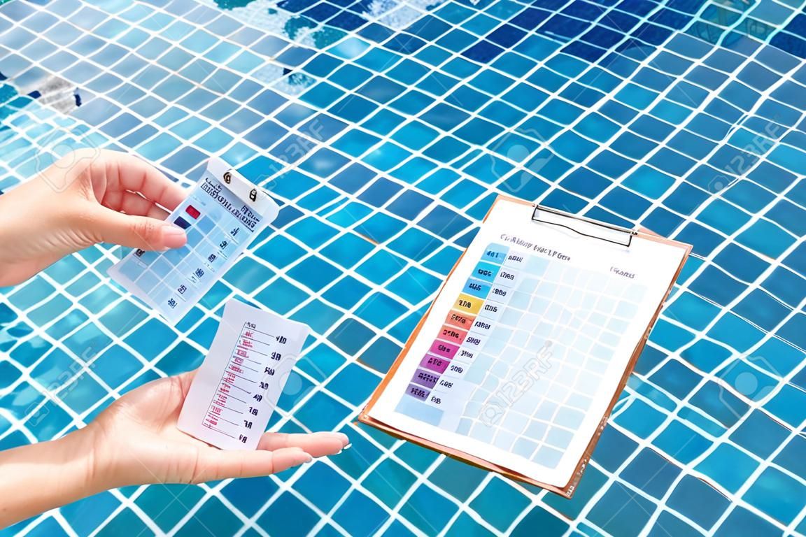 Female hand holding water tester and blank paper on wooden clipboard over clear swimming pool water background, pool water treatment checklist