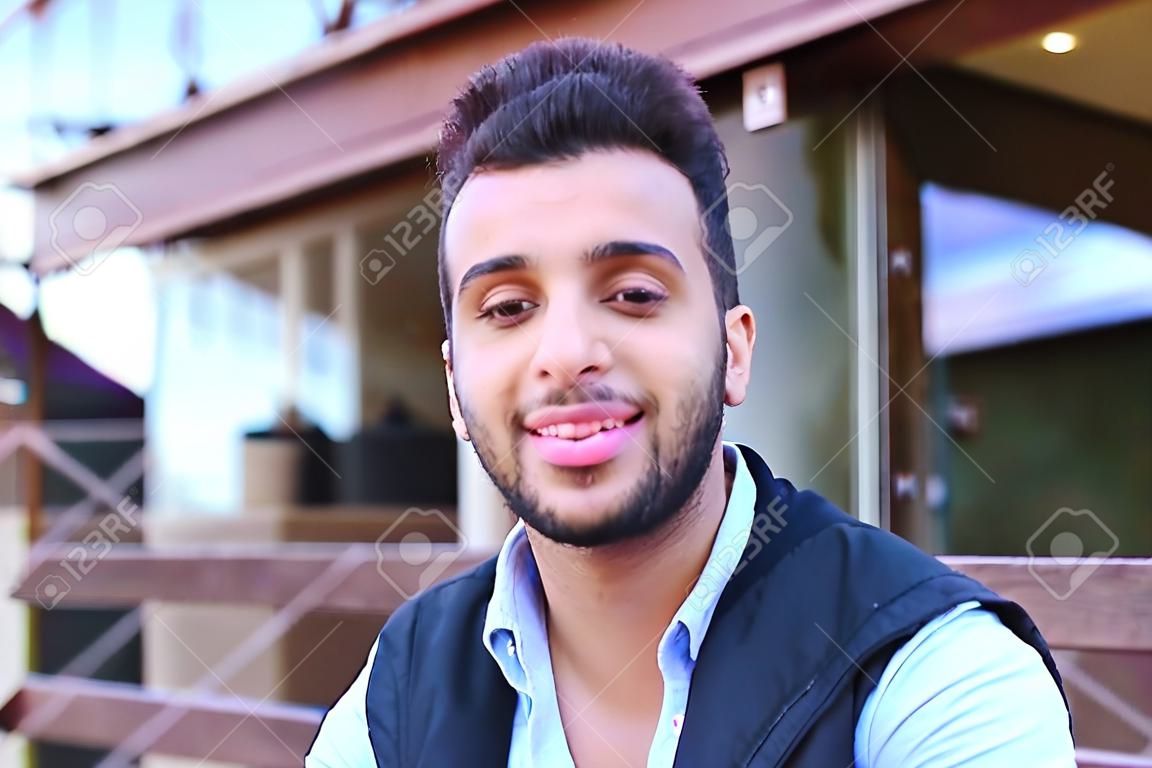 Portrait of beautiful Arab guy. Happy young man smiling big smile and looking in camera removed for advertising toothpaste or shampoo, stands on background of restaurant outdoors. Concept male cosmetics advertising, well-groomed guy healthy teeth and smil
