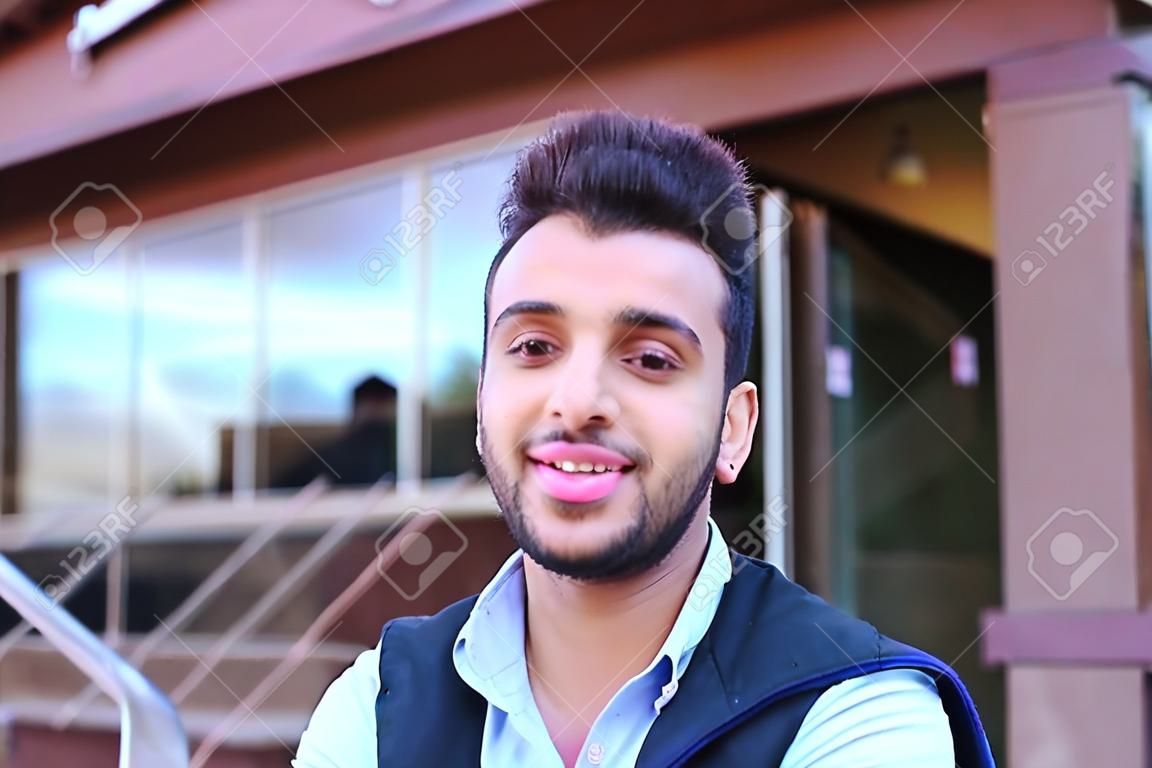 Portrait of beautiful Arab guy. Happy young man smiling big smile and looking in camera removed for advertising toothpaste or shampoo, stands on background of restaurant outdoors. Concept male cosmetics advertising, well-groomed guy healthy teeth and smil
