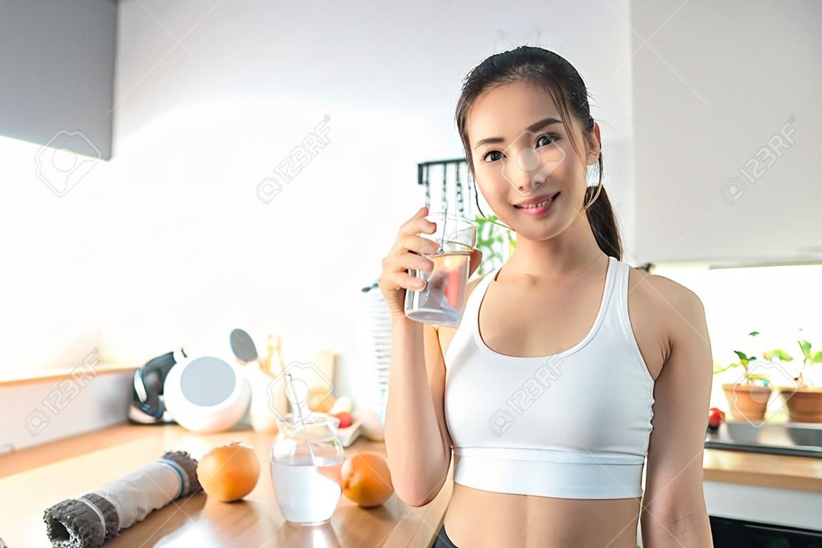 Portret van Aziatische vrouw in sportkleding drinken water na oefening thuis. Jonge dorstig actieve sport meisje neemt een slokje mineraal natuurlijk in de beker na de training en kijk naar camera in de keuken in huis.