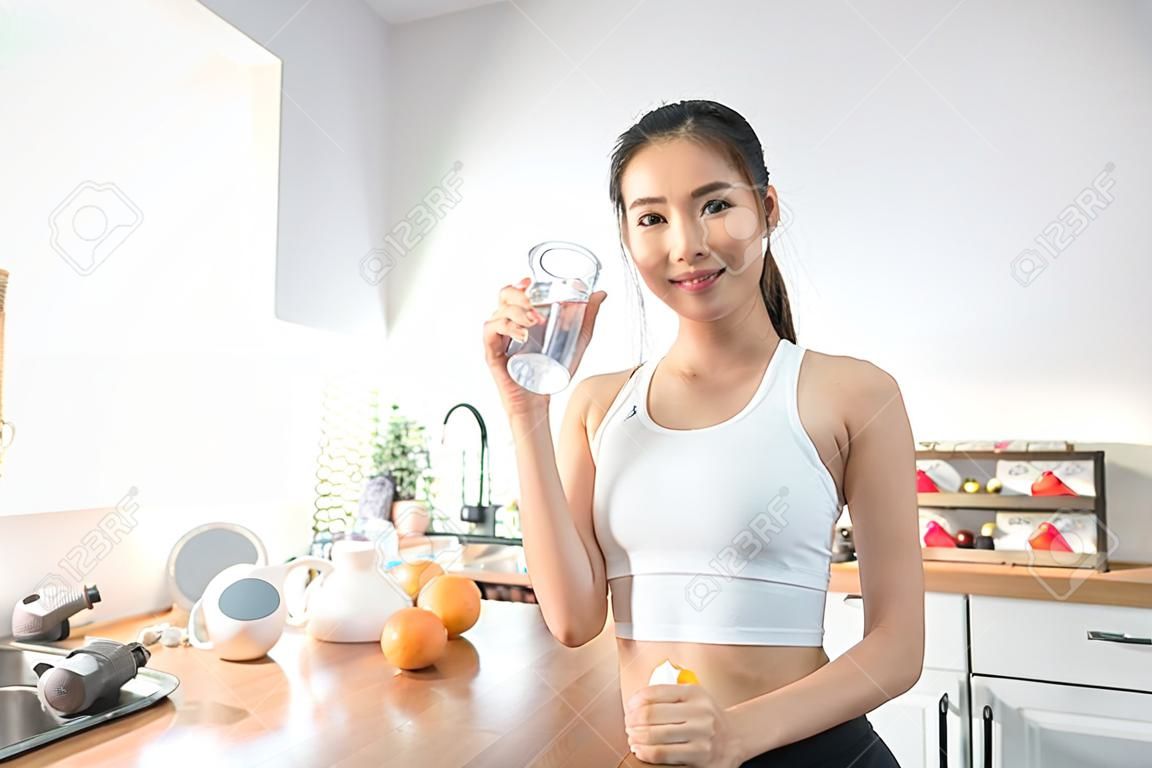Portret van Aziatische vrouw in sportkleding drinken water na oefening thuis. Jonge dorstig actieve sport meisje neemt een slokje mineraal natuurlijk in de beker na de training en kijk naar camera in de keuken in huis.