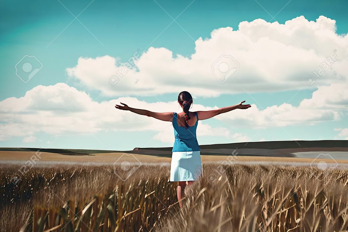 Vrouw staand in een maïsveld vreugdevol met haar rug naar de camera en armen uitgestrekt als ze kijkt naar het open platteland en bewolkte blauwe lucht