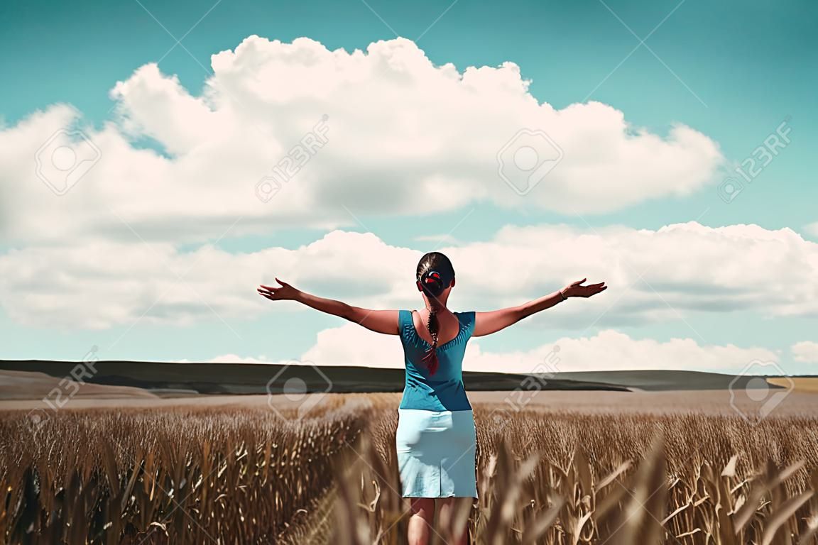 Mulher de pé em um campo de milho regozijando-se com as costas para a câmera e os braços estendidos enquanto ela enfrenta o campo aberto e céu azul nublado