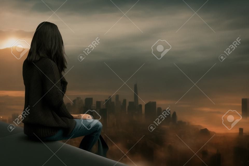 Woman wearing black sweater and facing away from camera while sitting in foreground above large metropolis