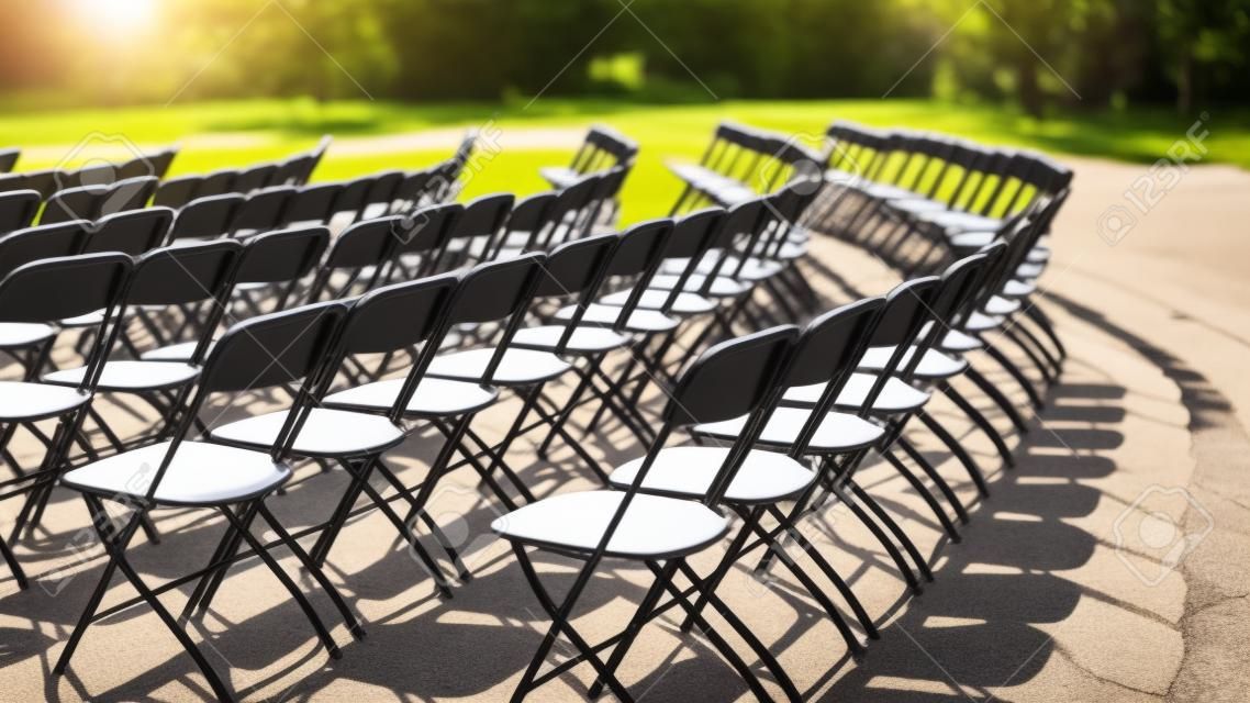 Empty seat rows of folding chairs on ground before a concert, parallel and rounded arranged, multiple black chairs on street on sunny day in park