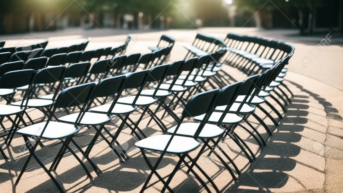Empty seat rows of folding chairs on ground before a concert, parallel and rounded arranged, multiple black chairs on street on sunny day in park