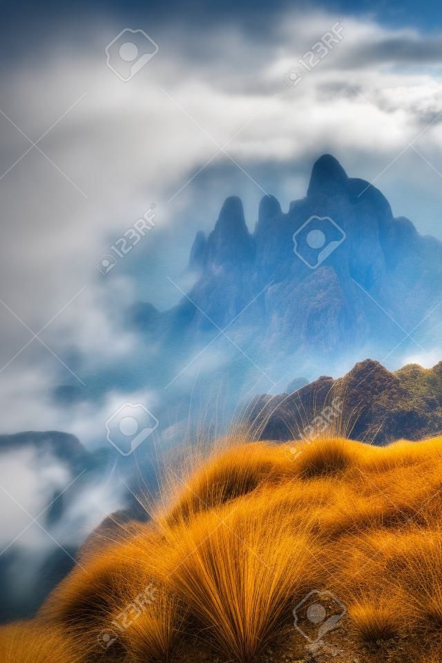 True tilt shift view of dry grass on rock in foreground and and distant mountains surrounded by low clouds in background, scenery hazy landscape of Thailand mountain peaks above clouds on moody day
