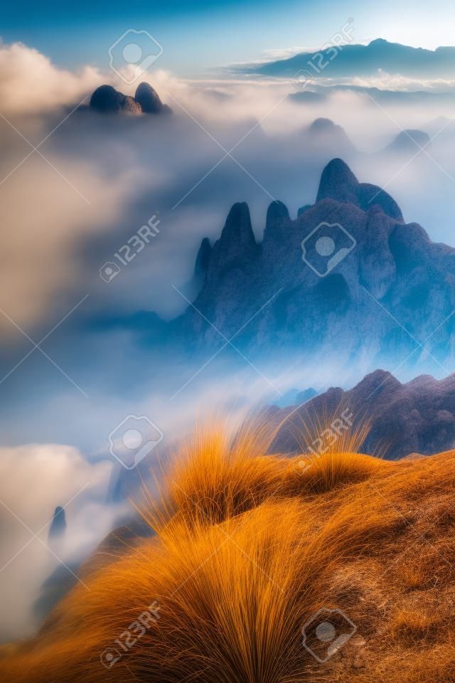 True tilt shift view of dry grass on rock in foreground and and distant mountains surrounded by low clouds in background, scenery hazy landscape of Thailand mountain peaks above clouds on moody day