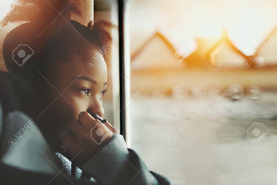 True tilt shift view of smiling black teenager girl near window traveling by ship, biracial young female is laughing and covering her mouth with hand while sitting inside of small cafe near water