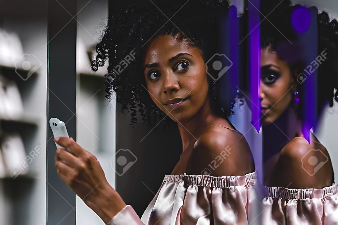 Une charmante jeune femme afro-américaine bouclée est appuyée contre le mur de verre noir tout en tenant le smartphone et en tapant un message à son amie; Femme brésilienne sur rue avec le téléphone portable
