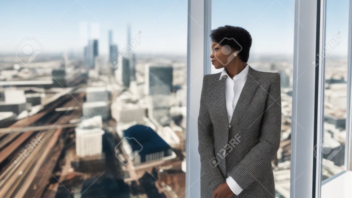 Zamyślona afroamerykańska przedsiębiorczyni w zamyśleniu patrzy na panoramę miasta za oknem, opierając się o ramę okna na najwyższym piętrze luksusowego wnętrza biura biznesowego