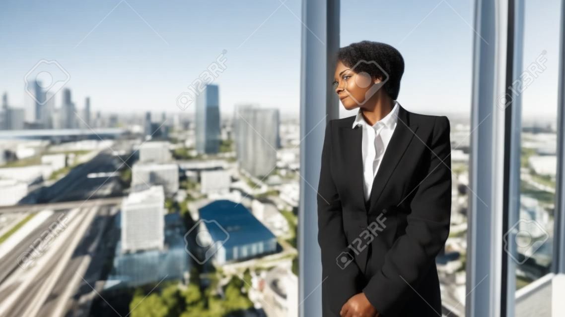Zamyślona afroamerykańska przedsiębiorczyni w zamyśleniu patrzy na panoramę miasta za oknem, opierając się o ramę okna na najwyższym piętrze luksusowego wnętrza biura biznesowego
