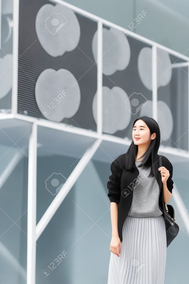 Sottile ragazza dai capelli neri in camicia bianca, gonna e borsa sportiva grigio su sfondo di ventilazione