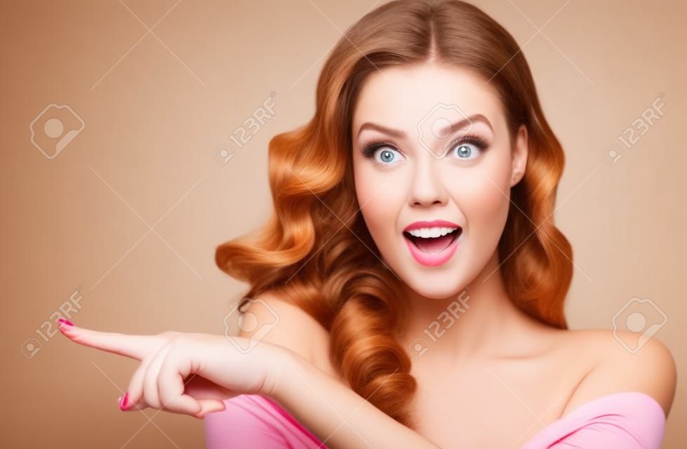 Une femme surprise montre un produit invisible. Belle fille aux cheveux bouclés pointant sur le côté. Expression du visage lumineux.