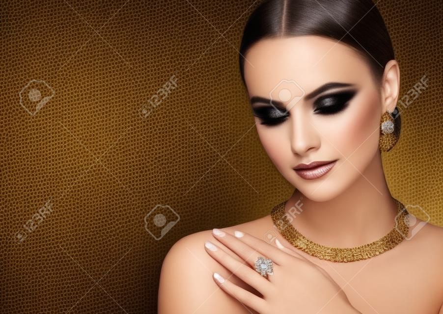 스모키 아이 메이크업 스타일의 예쁜 모델이 금박 보석 세트를 선보이고 있습니다. 귀걸이, 목걸이, 반지가 포함된 금박 보석 세트는 완벽하게 보이는 젊은 여성에게 옷을 입습니다.