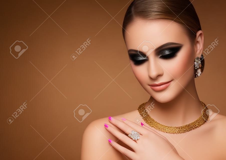 Bonita modelo con estilo de maquillaje de ojos ahumados está demostrando un conjunto de joyas doradas. El conjunto de joyas doradas que contiene aretes, collar y anillo está vestido por una mujer joven de aspecto perfecto.
