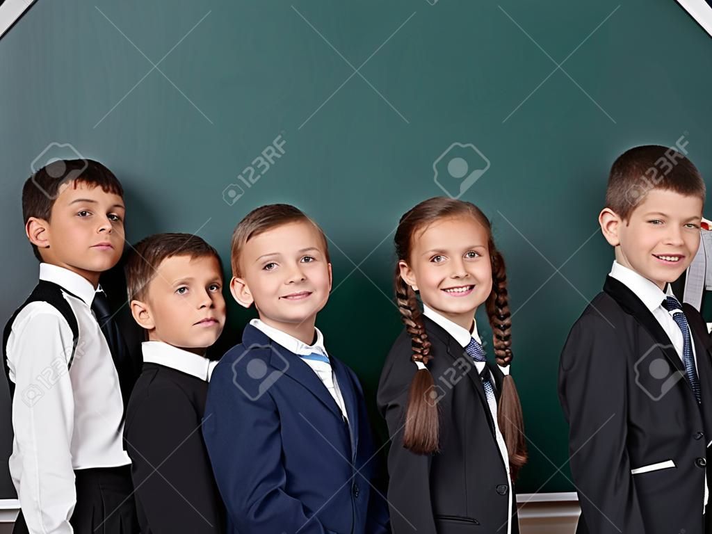 Niño de escuela primaria cerca de pizarra en blanco de fondo, vestido de traje negro clásico, grupo de alumnos, concepto de educación