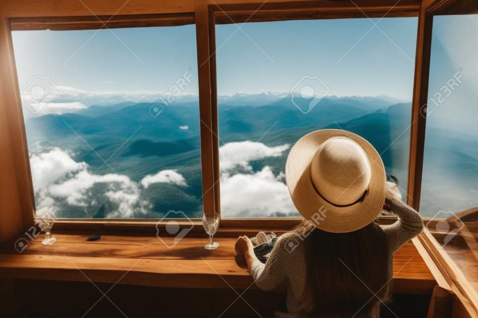 Stylish hipster girl in hat looking on top of mountains through big window in old cabin. Young woman traveler relaxing  in summer mountains. Travel and wanderlust. Amazing moment