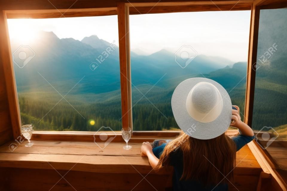 Stijlvolle hipster meisje in hoed kijken op de top van de bergen door grote raam in oude cabine. Jonge vrouw reiziger ontspannen in de zomer bergen. Reizen en wandellust. Verbazingwekkend moment